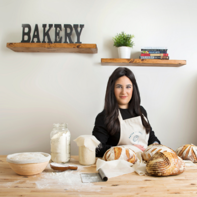 Woman with dark hair in apron smiling at baking table with sourdough bread ingredients