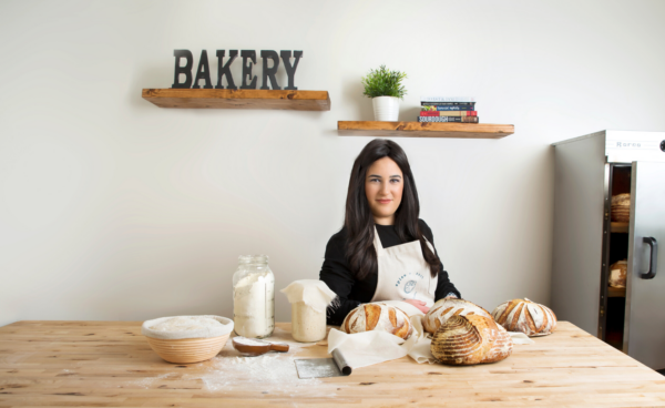 Woman with dark hair in apron smiling at baking table with sourdough bread ingredients