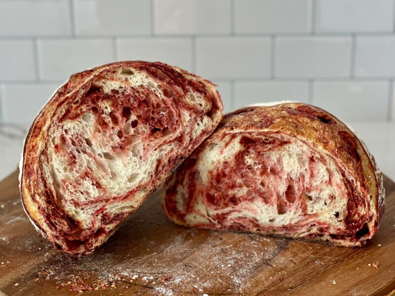 Sourdough bread with a vibrant red beet swirl, cut in half and displayed on a wooden cutting board.