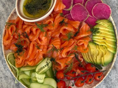 Homemade gravlax recipe—thin‑sliced cured salmon with dill on a platter with cucumber ribbons, avocado, watermelon radish, cherry tomatoes, and dill dressing.