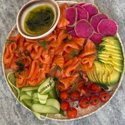 Homemade gravlax recipe—thin‑sliced cured salmon with dill on a platter with cucumber ribbons, avocado, watermelon radish, cherry tomatoes, and dill dressing.